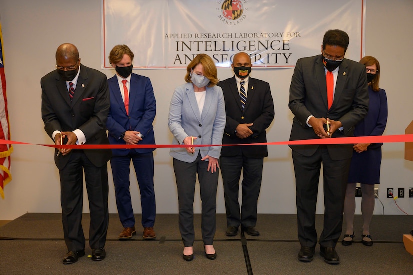 Three people cut  a red ribbon while others watch.