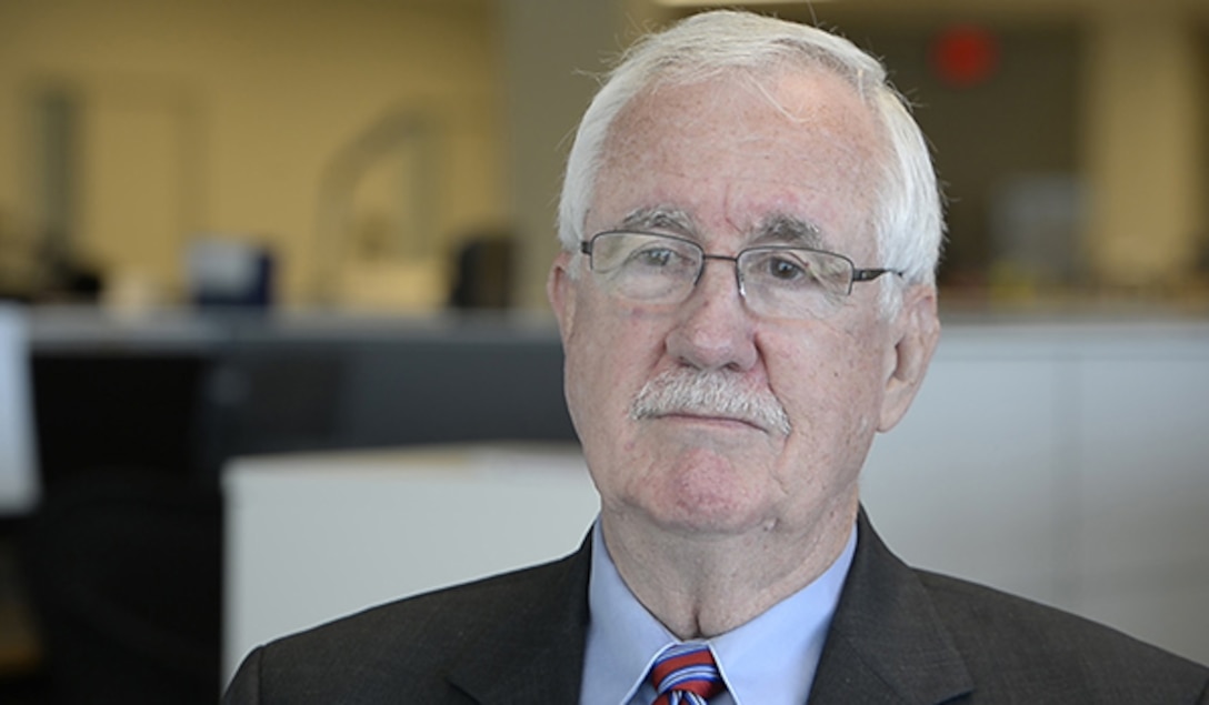 An older man in a suit and glasses looks past a camera.