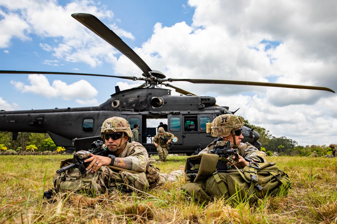 Soldiers pull security on the ground with a helicopter in the background.