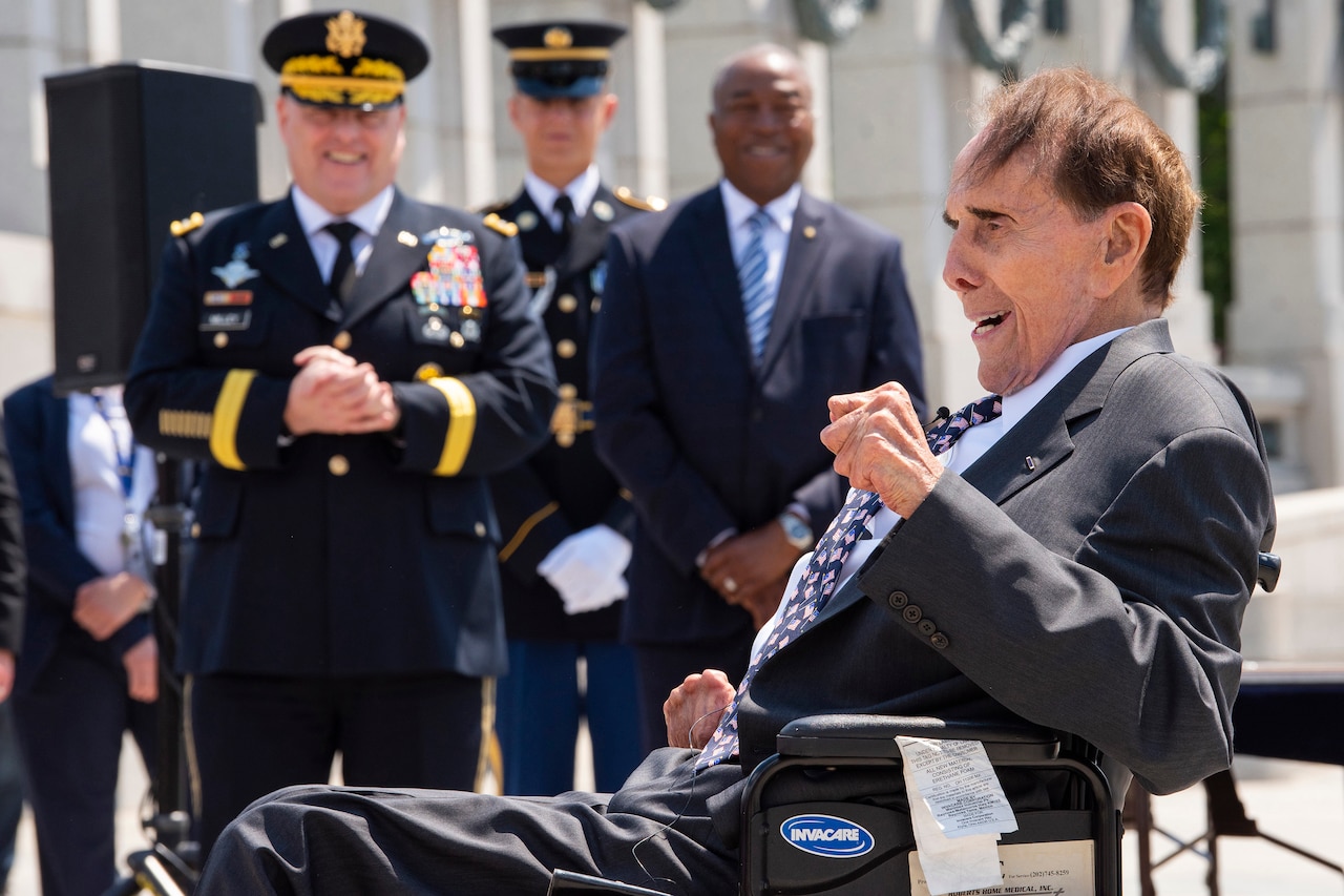 A man wearing a suit and seated in a wheelchair smiles as he speaks.