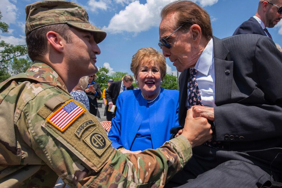 Soldier in camouflage uniform and man wearing a suit clench hands as a woman in a blue dress stands between them.