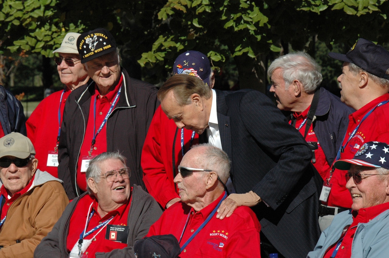 Man wearing a suit stands among veterans wearing red shirts as he bends to speak with one who is seated.