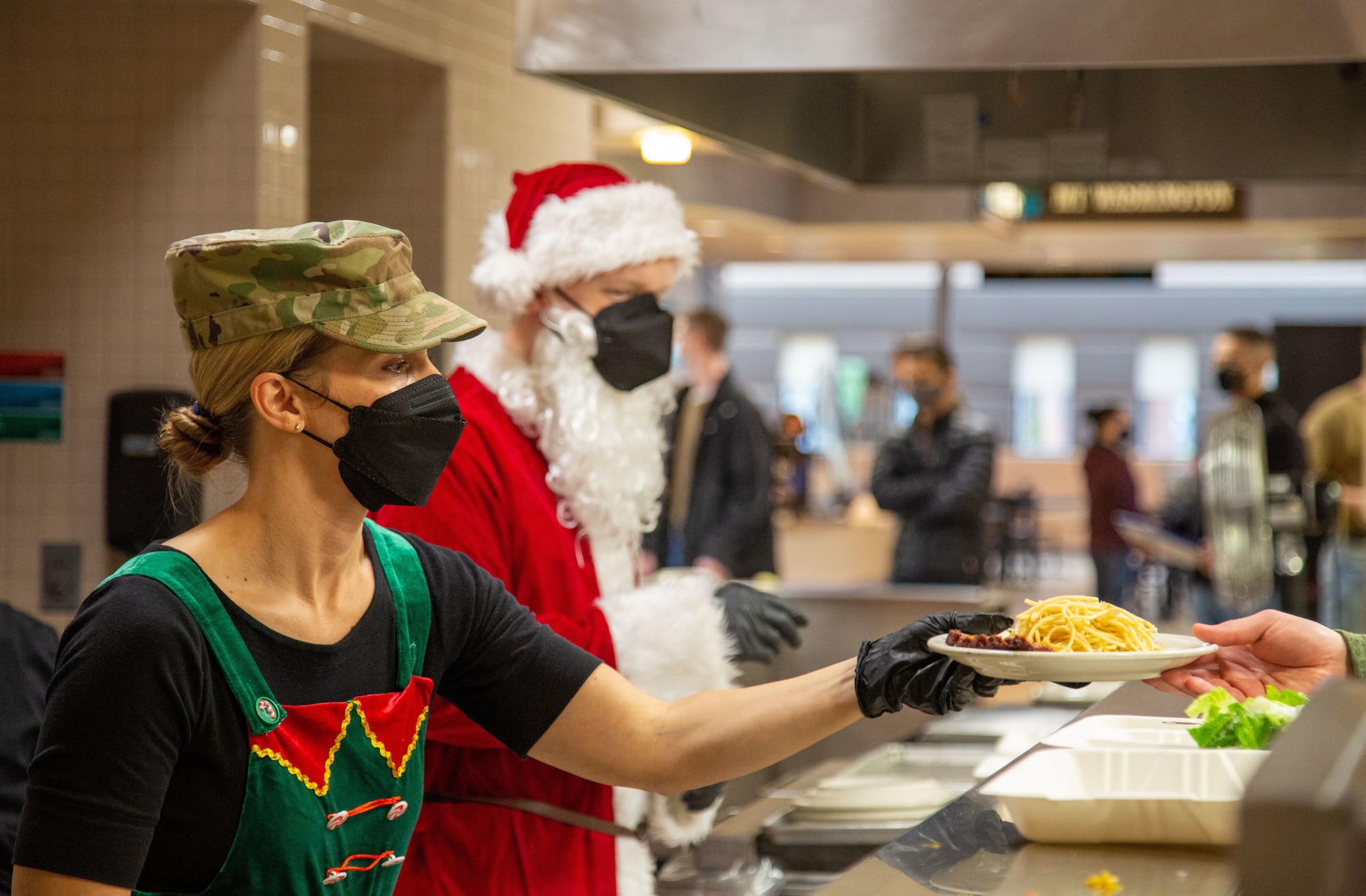 An elf with a military hat and a Santa serve food to patrons in dining facility.