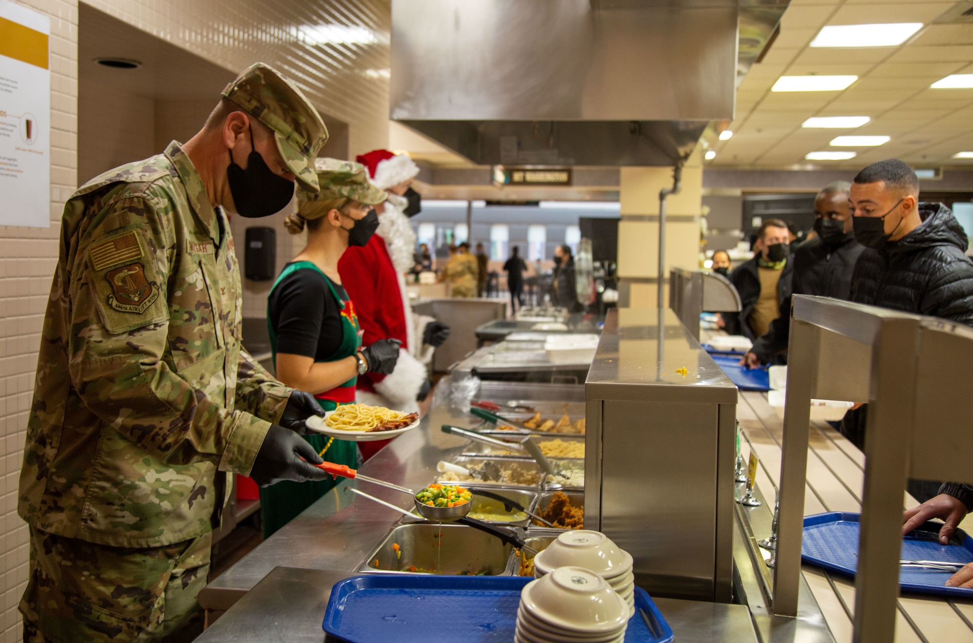 A military leader, an elf and a Santa serve food to patrons in dining facility.