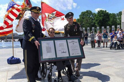 Two soldiers flank a man in a wheelchair as they display a set of framed proclamations.