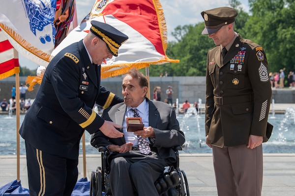 Two soldiers in uniform flank a man in a wheelchair while speaking with him.