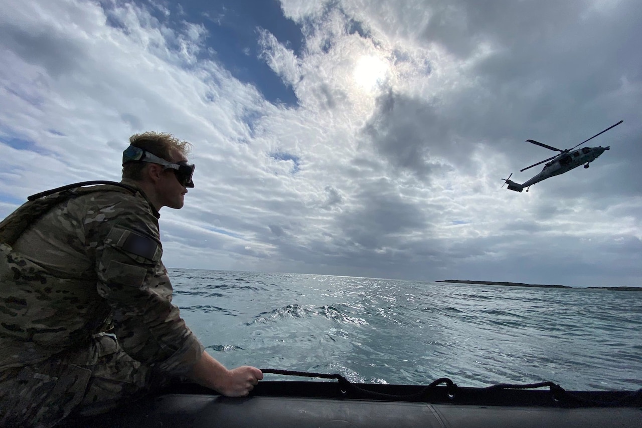 A soldier rides in a small boat as a helicopter flies nearby.