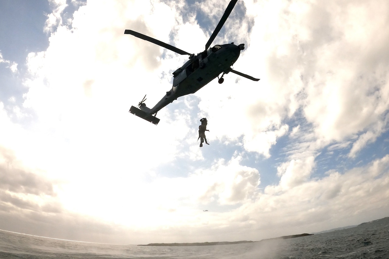 Soldiers attached to a rope hold onto a hovering helicopter above water.