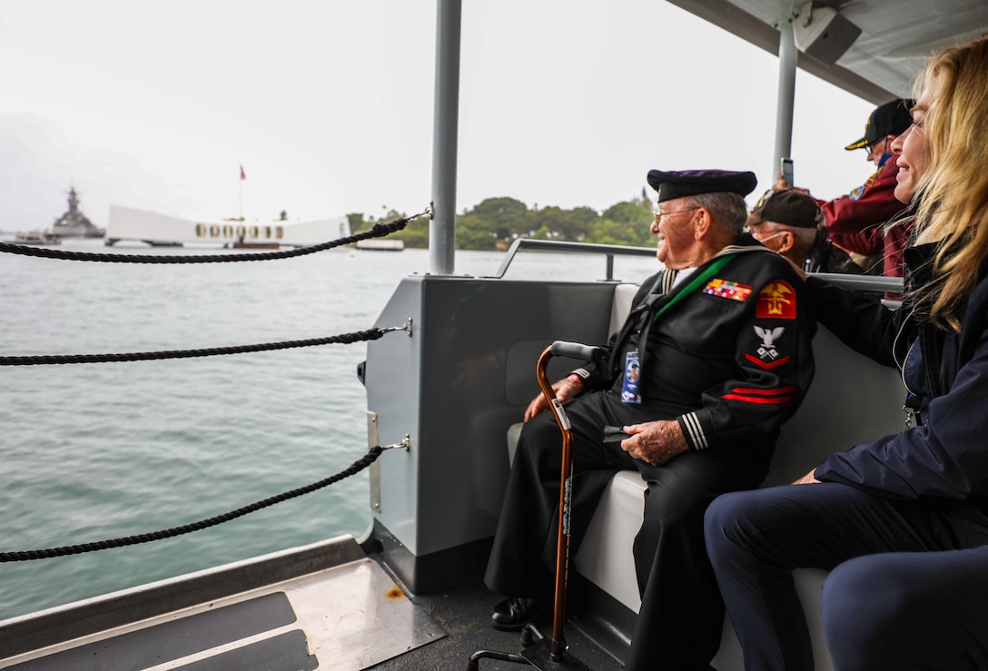 A veteran in a sailor's uniform and others on a boat look at a memorial in the distance.