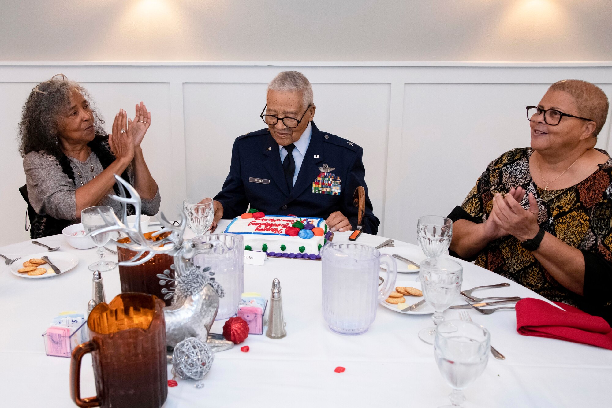 Man in Air Force uniform looking at a birthday cake. His daughters on either side of him.