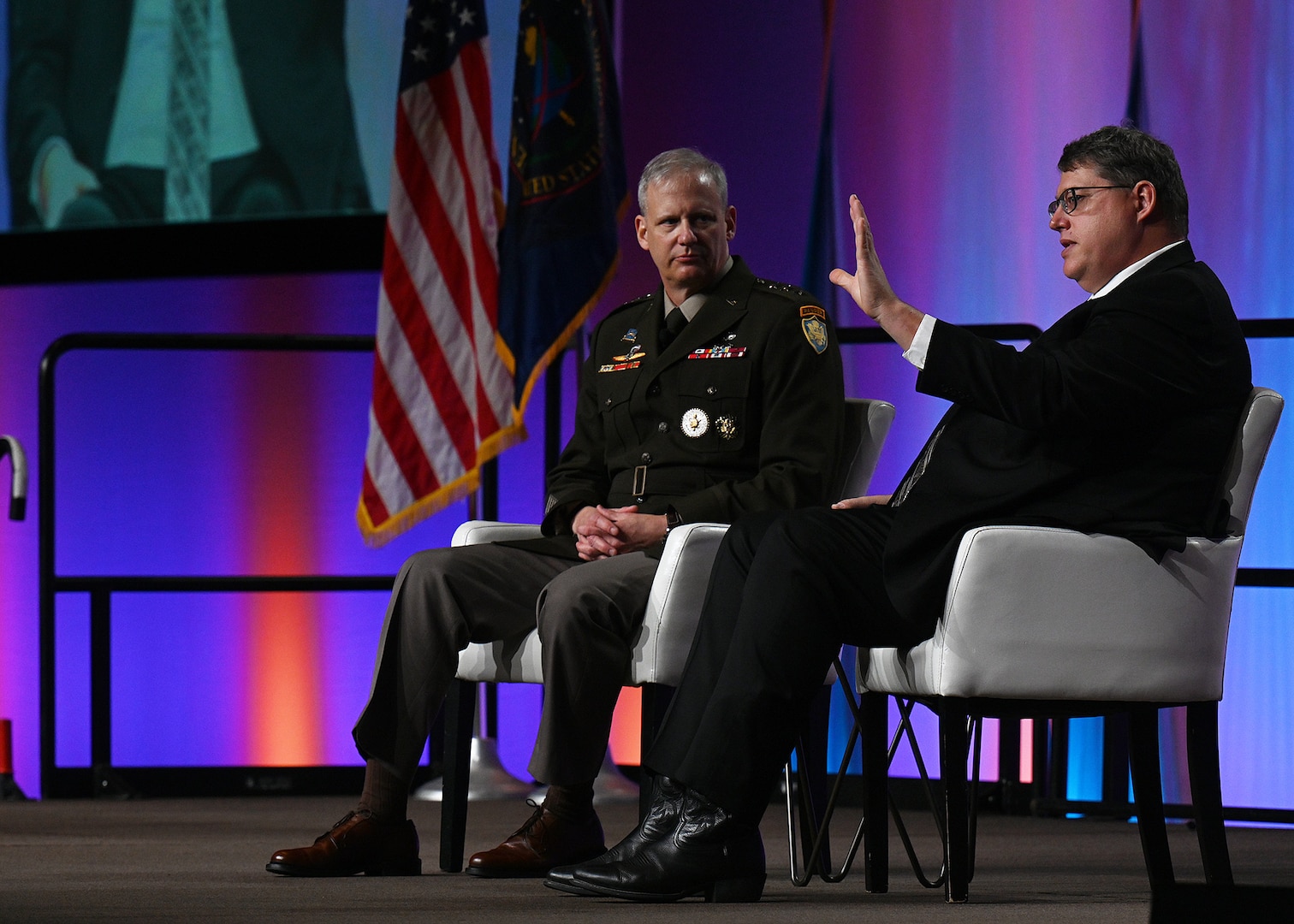 Image of two men seated on a stage talking toward an audience.
