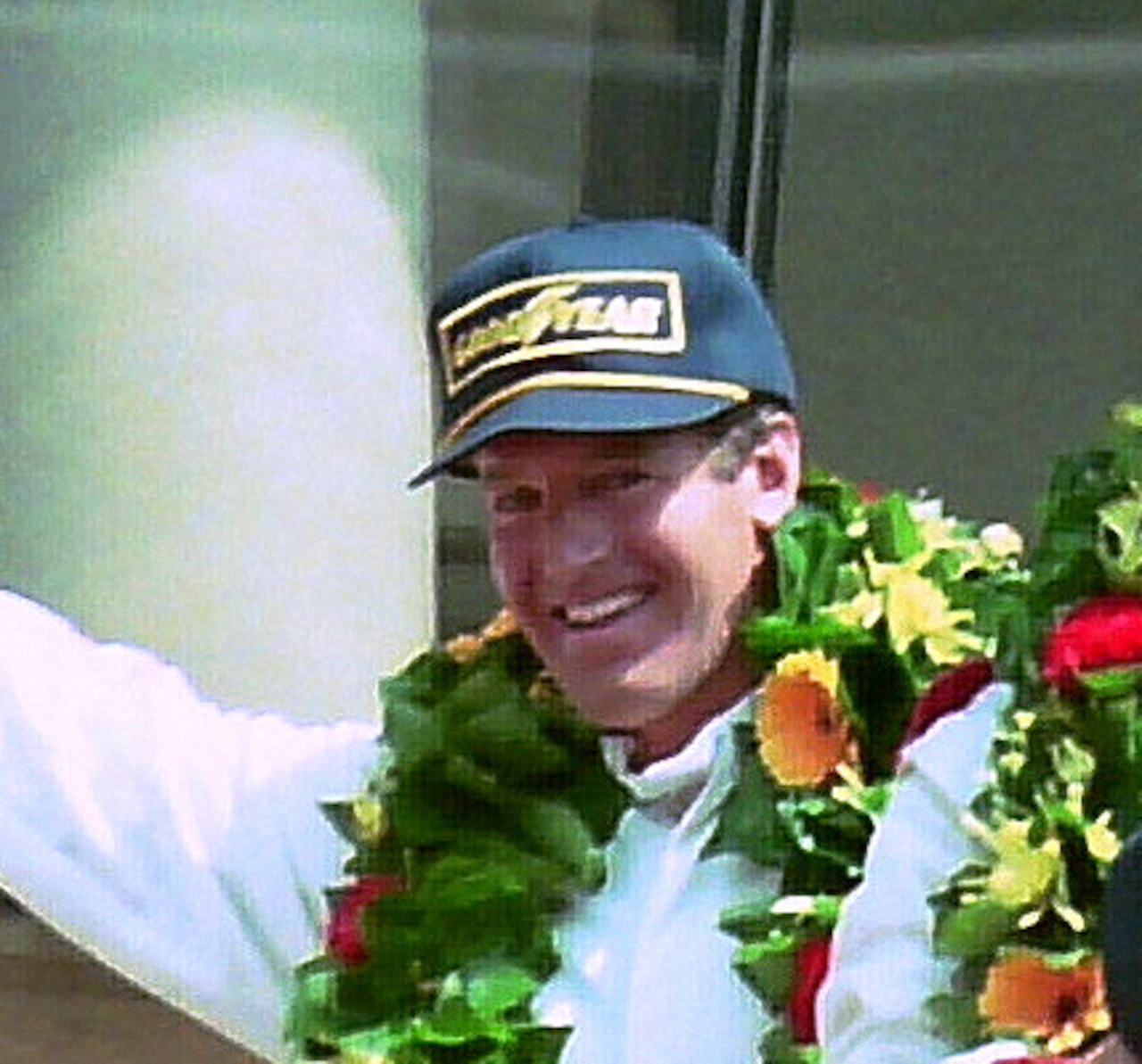 A man, wreathed in flowers, smiles.