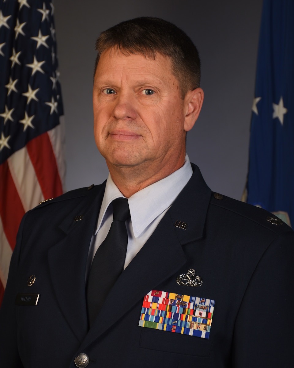 A man poses in Air Force service dress with an American flag behind his right shoulder, and an Air Force flag behind his left shoulder in front of a neutral gray background.