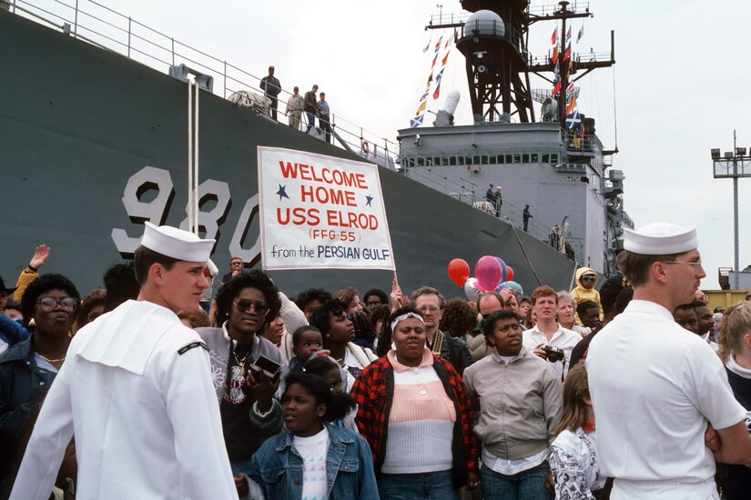 Dozens of people stand in front of a large ship with a sign that reads "Welcome home USS Elrod." Two sailors stand in front of them.