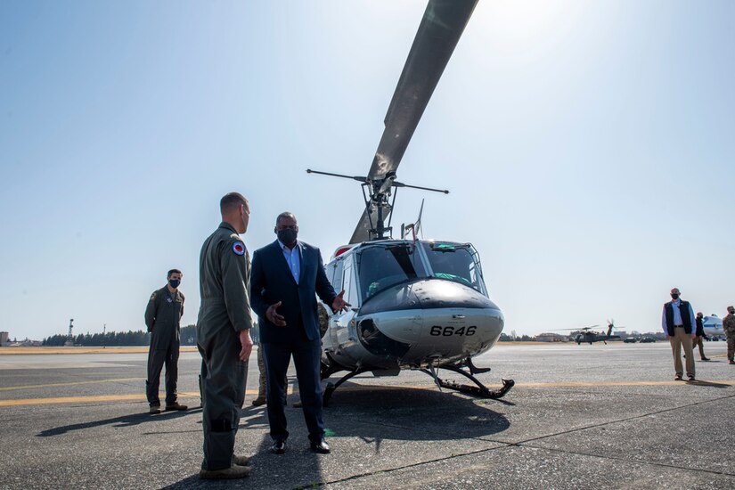 In the foreground, two men stand near a helicopter. Another man is in the background.