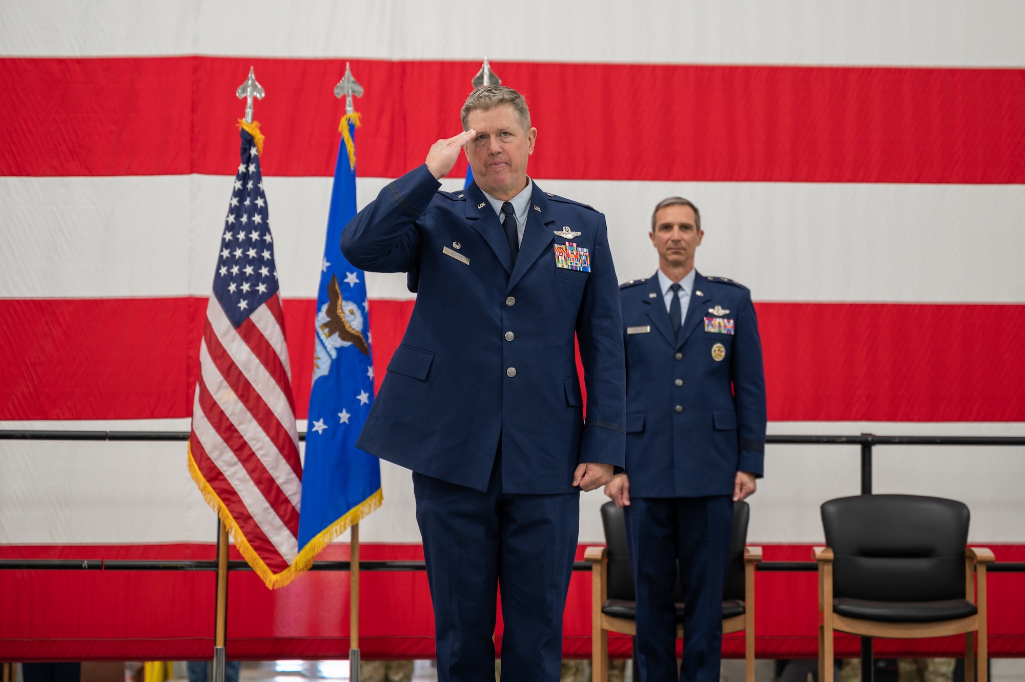 Two men in U.S. Air force service dress stand in front of a large American flag. The one in the foreground is rendering a salute.