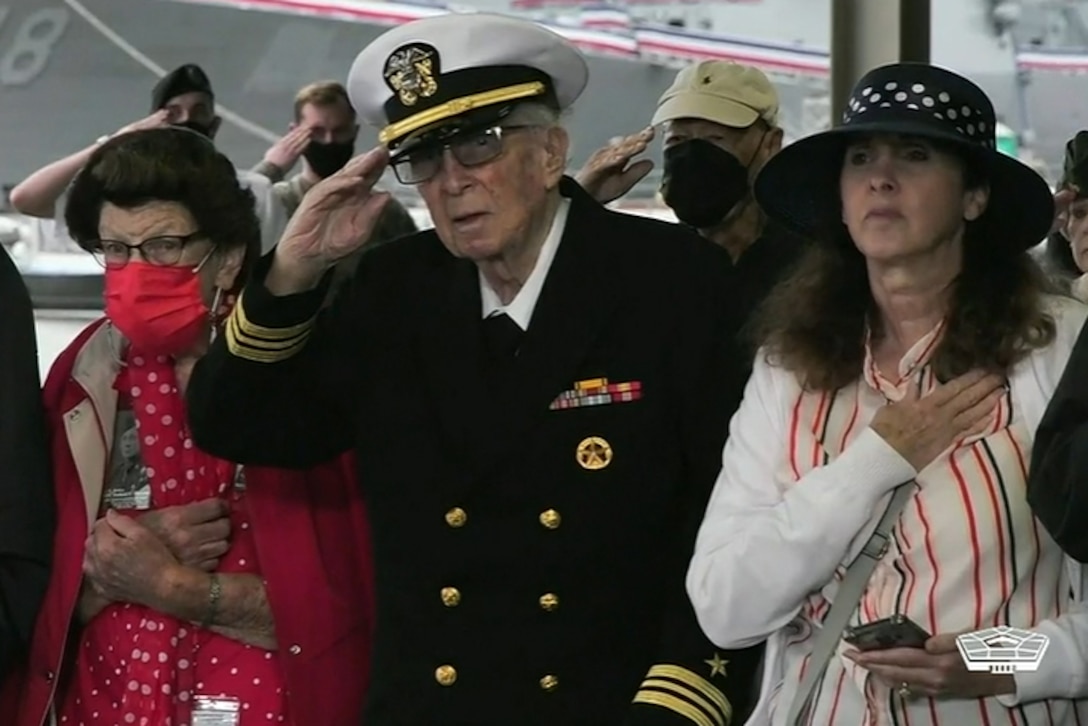 An elderly man in uniform salutes, as do others gathered around him.