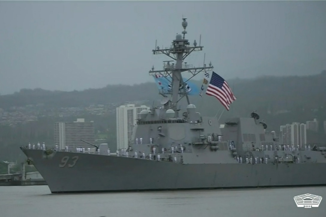 People stand along the deck of a ship as a U.S. flag whips in the wind.