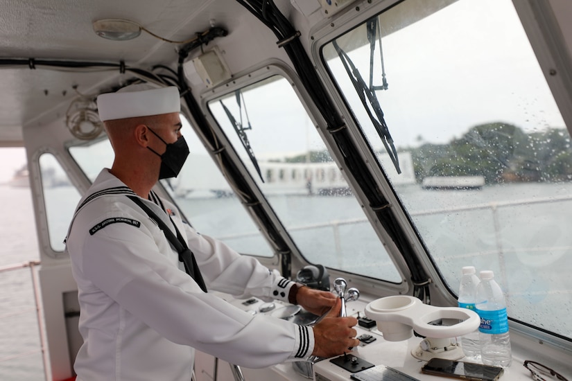 A sailor in dress whites and a face mask stands at the controls of a boat on the water, a memorial visible from the windows.