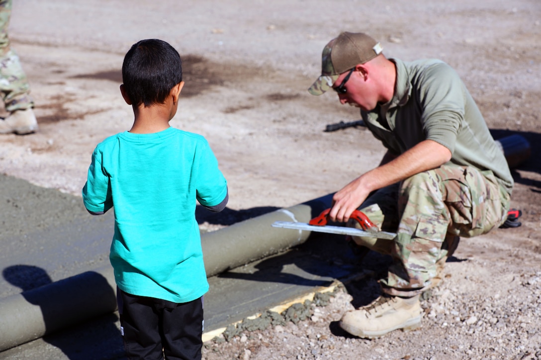 An Army soldier holding a trowl kneels down to smooth concrete while an Afghan child looks on.