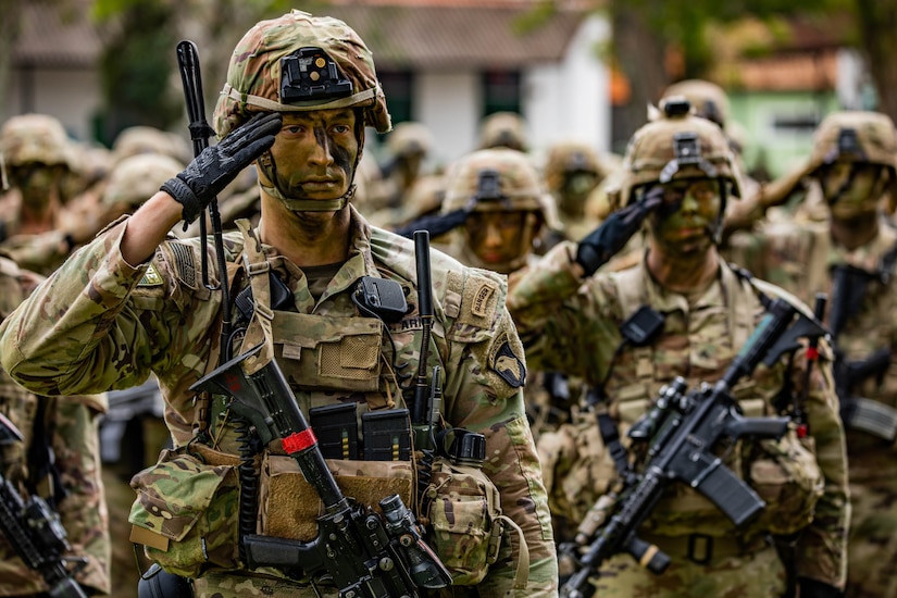 Soldiers in camouflage paint salute during a ceremony.
