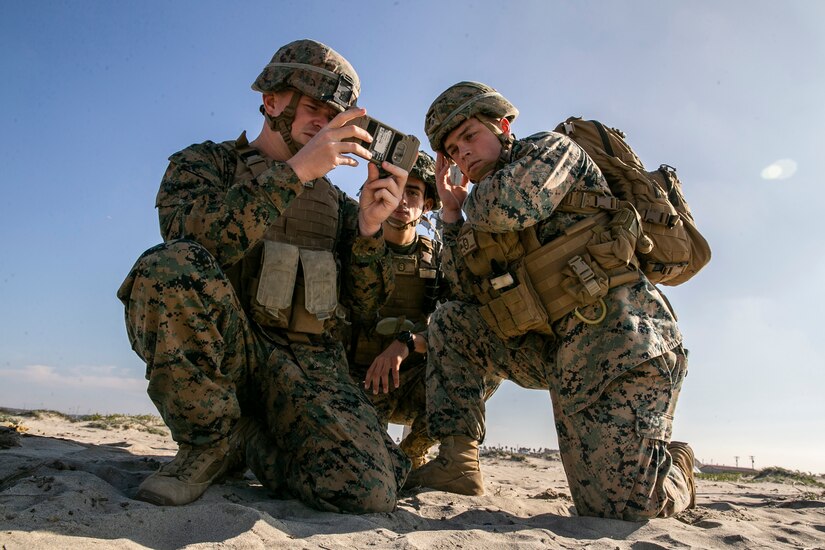 Three Marines kneel on the sand and look at a device.