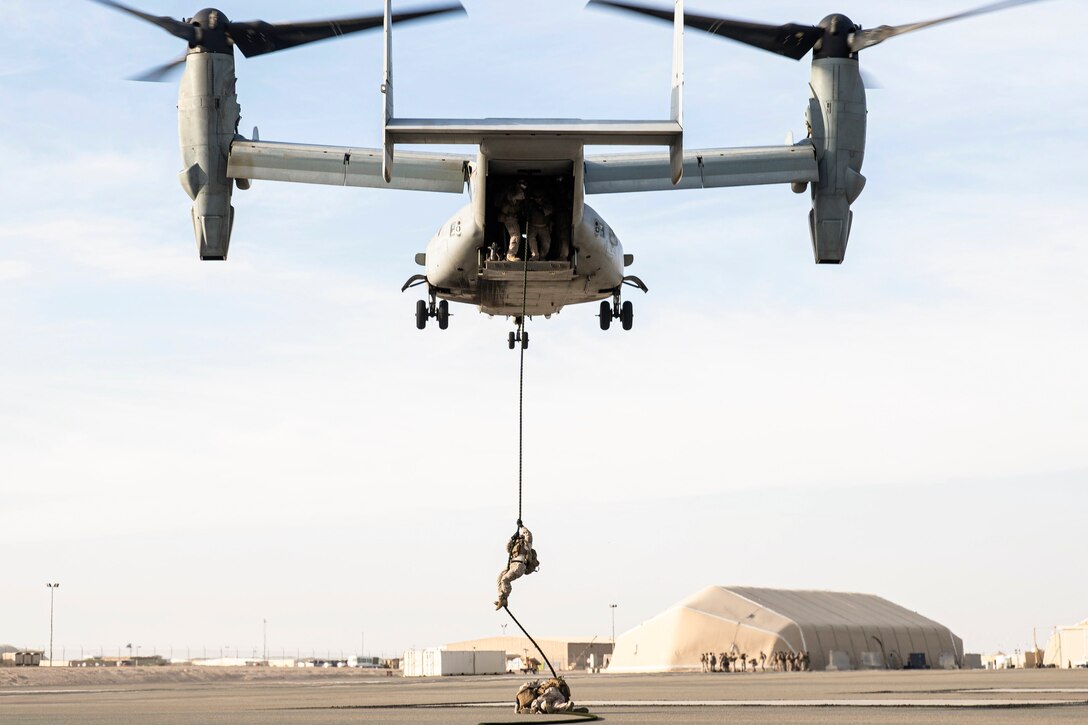 A Marine holds on to a rope attached to an airborne helicopter as fellow Marines watch.