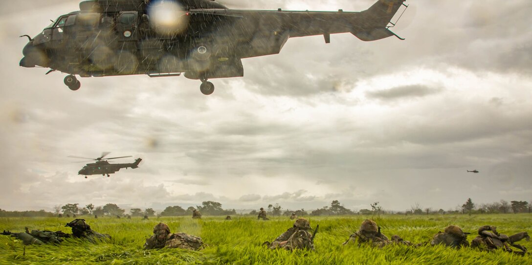 U.S. and Brazilian soldiers lay in the grass as a helicopter takes off.