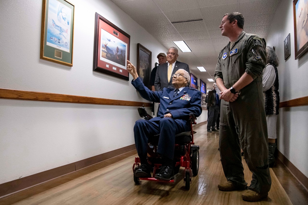 A man in a wheelchair points at a picture on the wall as others stand near him.