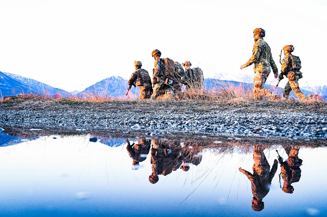 Soldiers walking across a field are reflected in a body of water.