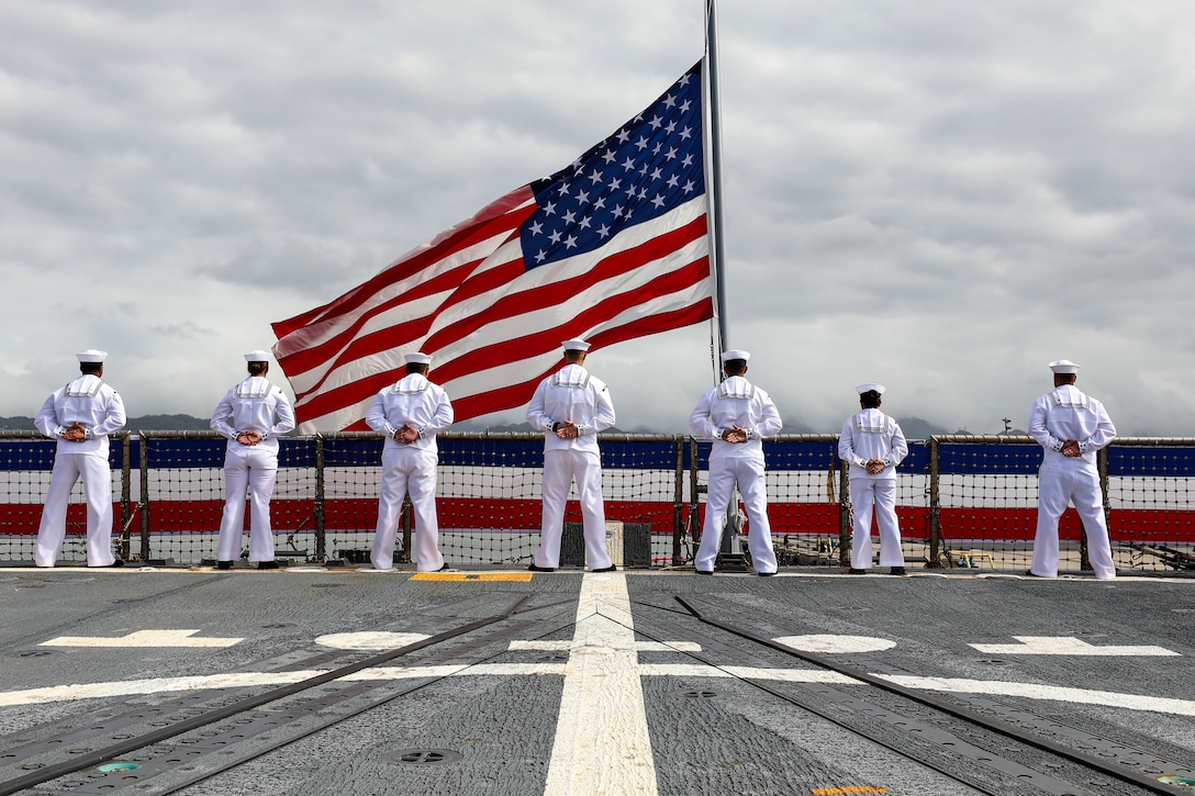 A row of sailors stand with their backs to the camera along the rail of a ship with the American flag flying.