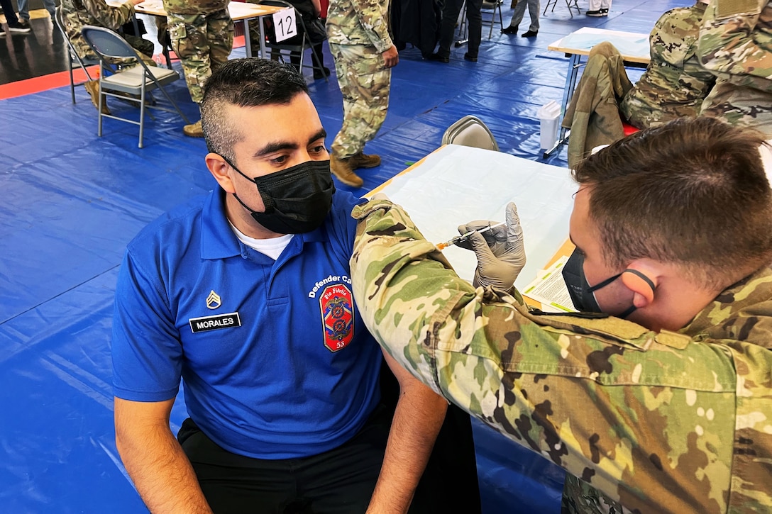 A soldier wearing a face mask and gloves gives a COVID-19 vaccine to a man sitting and wearing a face mask.