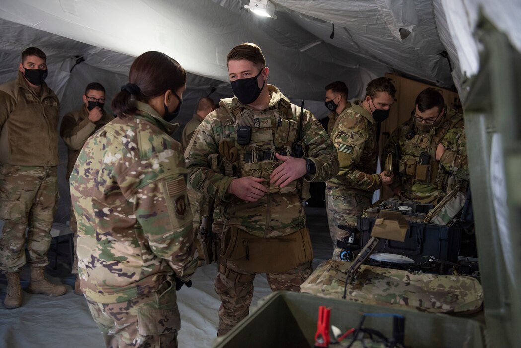 Two Airmen have a discussion in a tent about a drone that is displayed on a table, with a group of Airmen listening in behind them