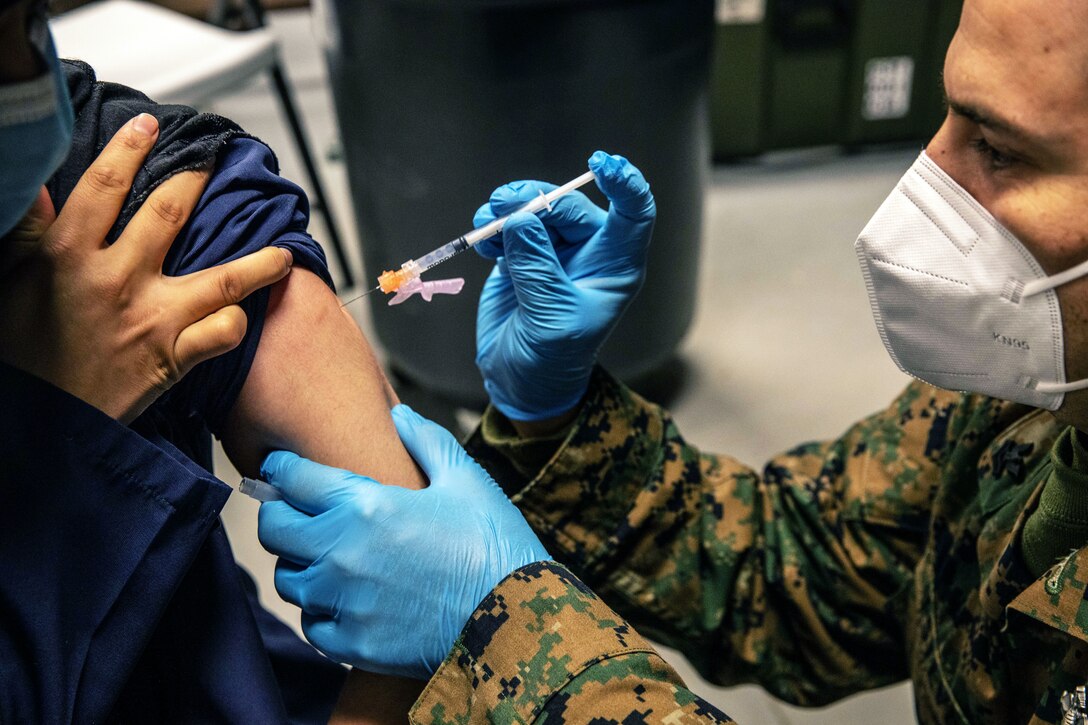 A sailor wearing face mask and gloves holds a syringe while administering a COVID-19 vaccine.