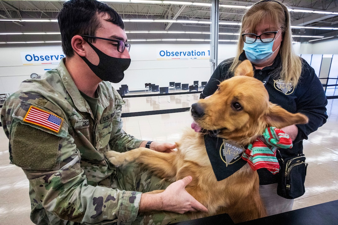 A soldier wearing a mask pets a therapy dog while woman wearing a mask stands in the back and looks on.