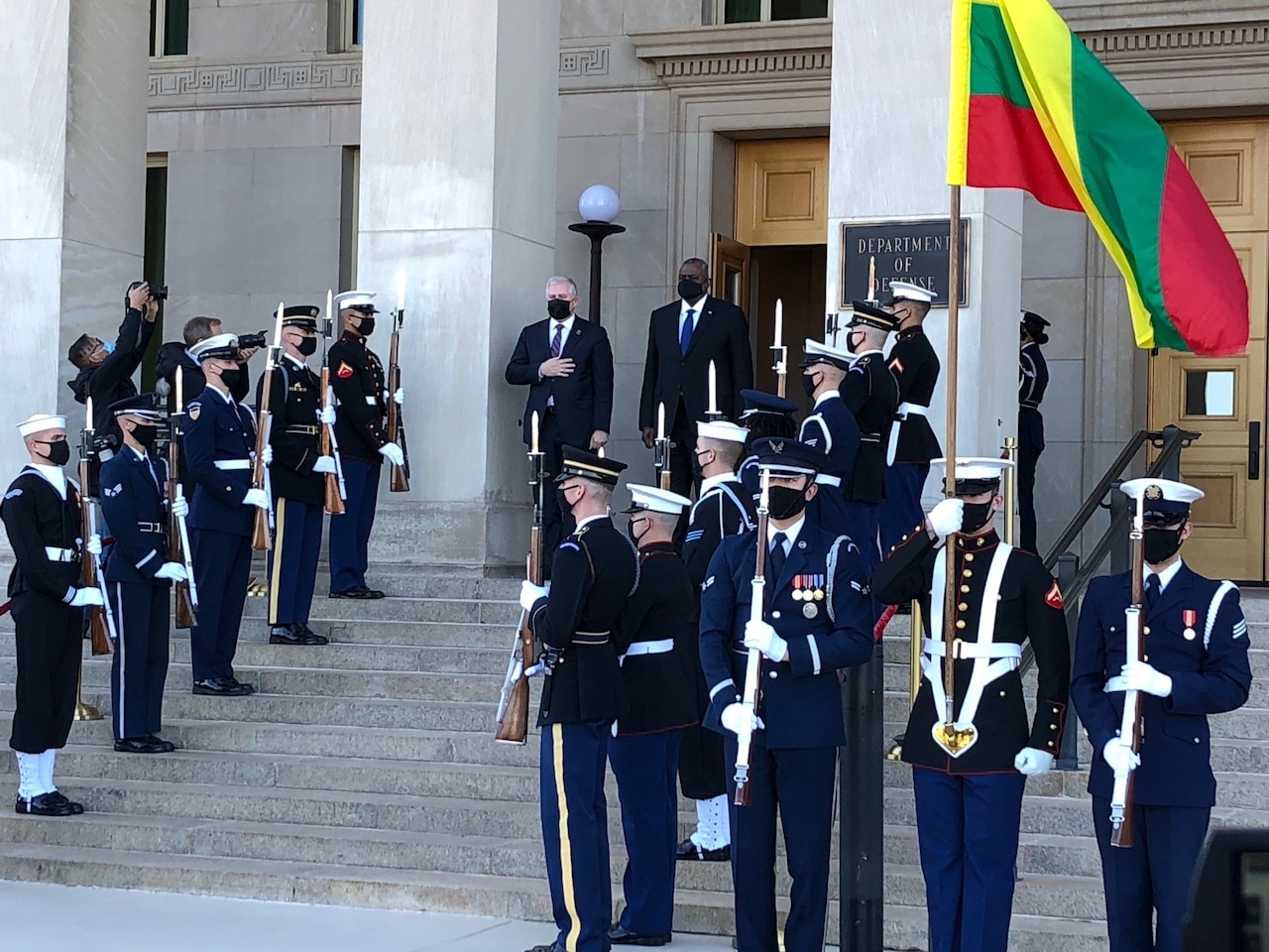 Two men stand on the top step outside a building between rows of service members. The sign on the building indicates that it is the Department of Defense.