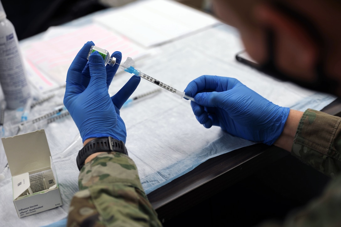 A soldier wearing gloves holds a syringe inserted into a vial.