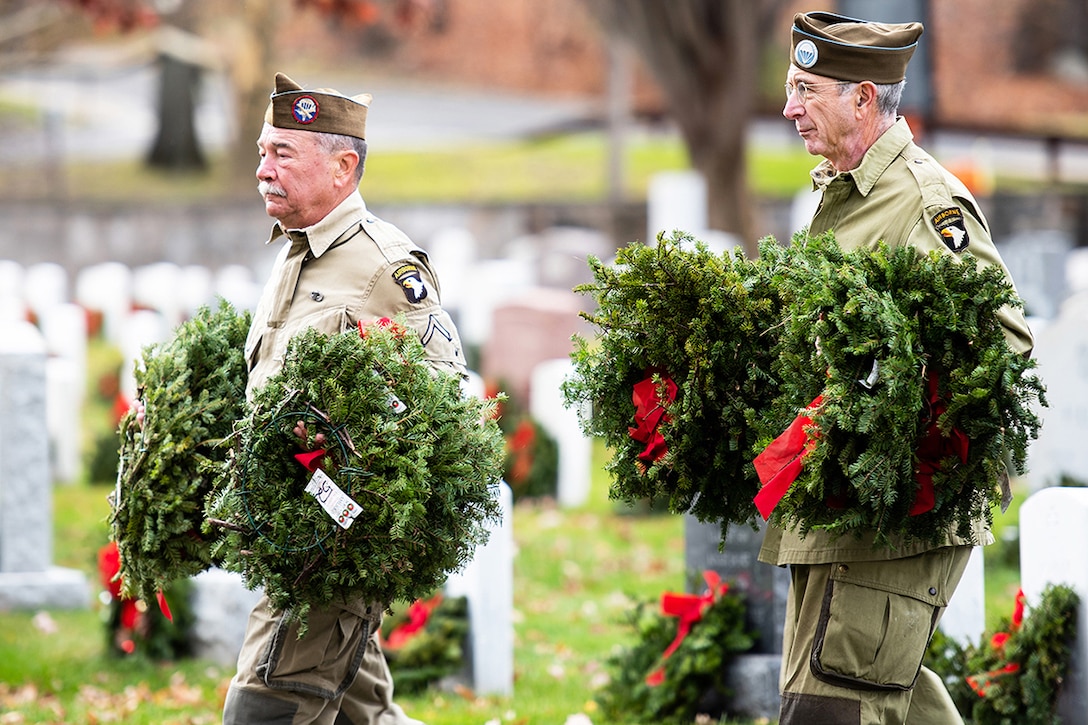 Two veterans carry wreaths in a cemetery.