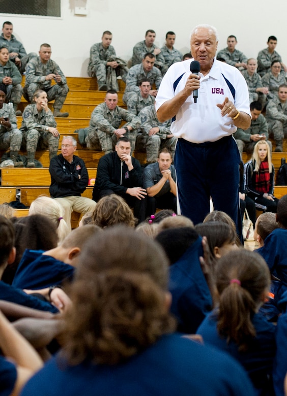 A man speaks to an audience of children while airmen look on from the background.