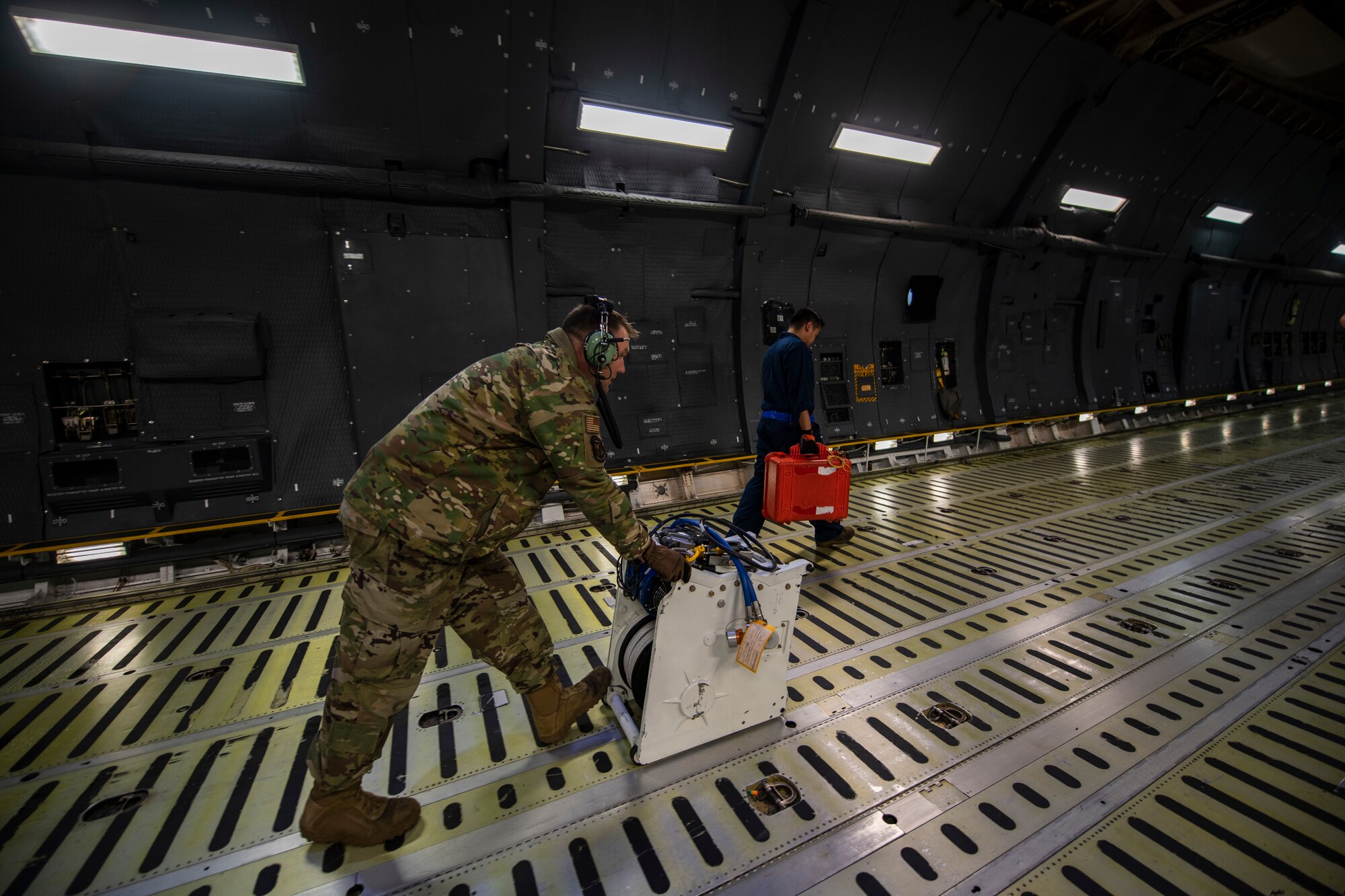 A man in military uniform pushes a piece of machinery with rope that is used to haul heavy objects on to a very large military aircraft.