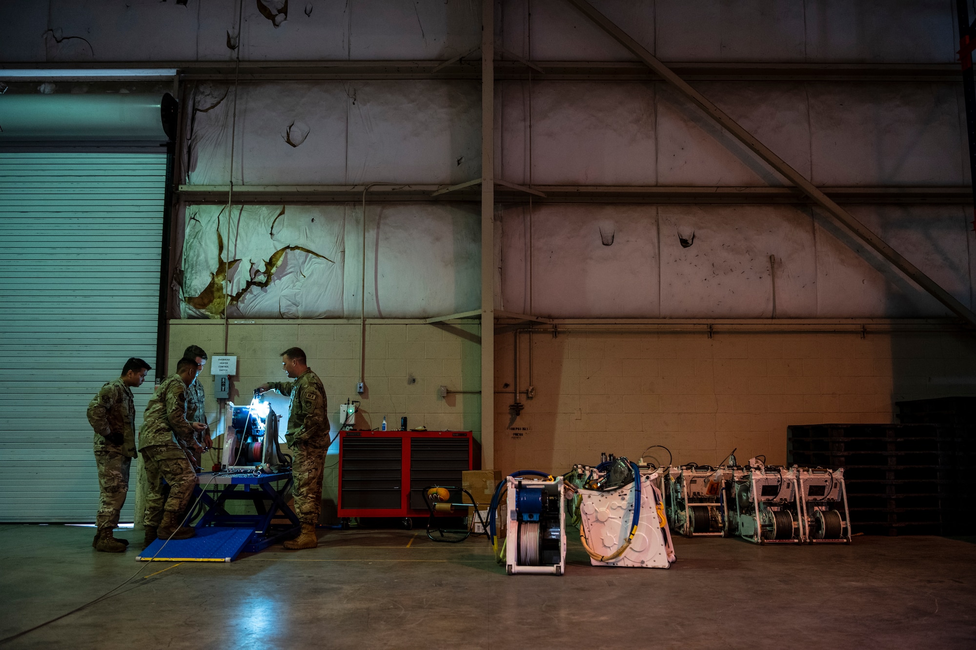 A wide show of a warehouse with men in military uniform working on a piece of machinery with rope on it.