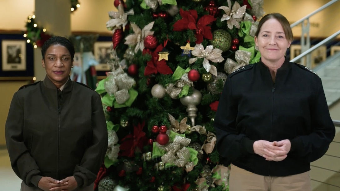 A Black woman and white woman, both in black jackets, stand in front of a Christmas tree decorated with ornaments.