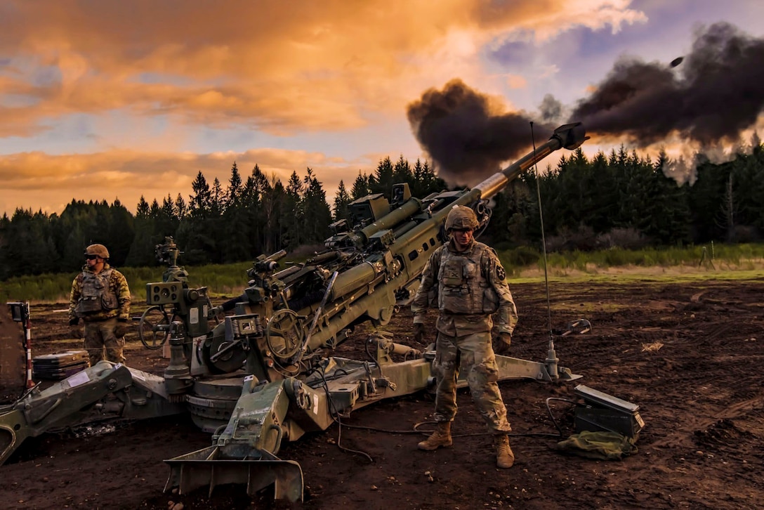 Two soldiers stand next to a military weapon as it fires.