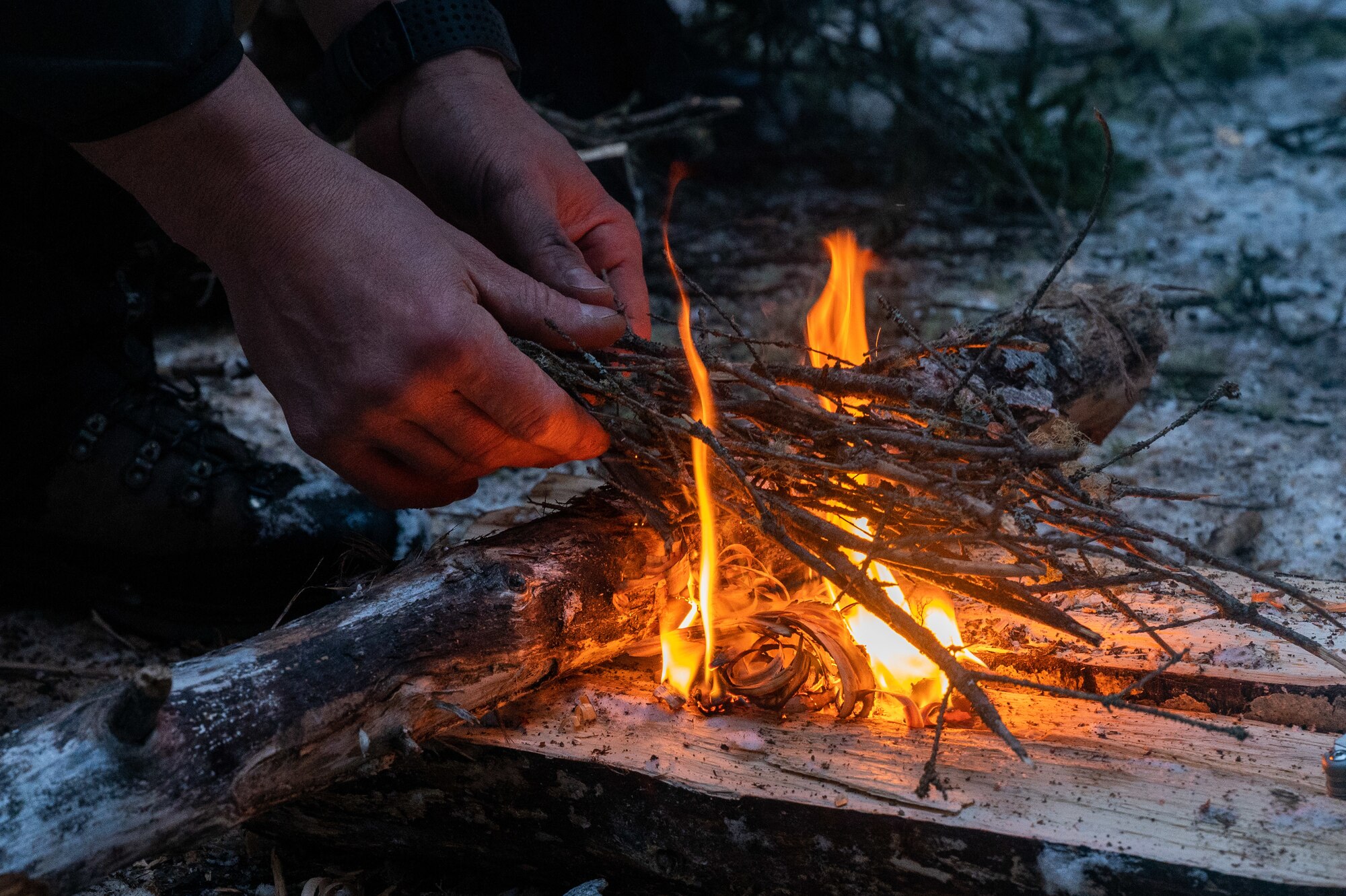 Hands place twigs on a fire on top of a log resting on the ground.