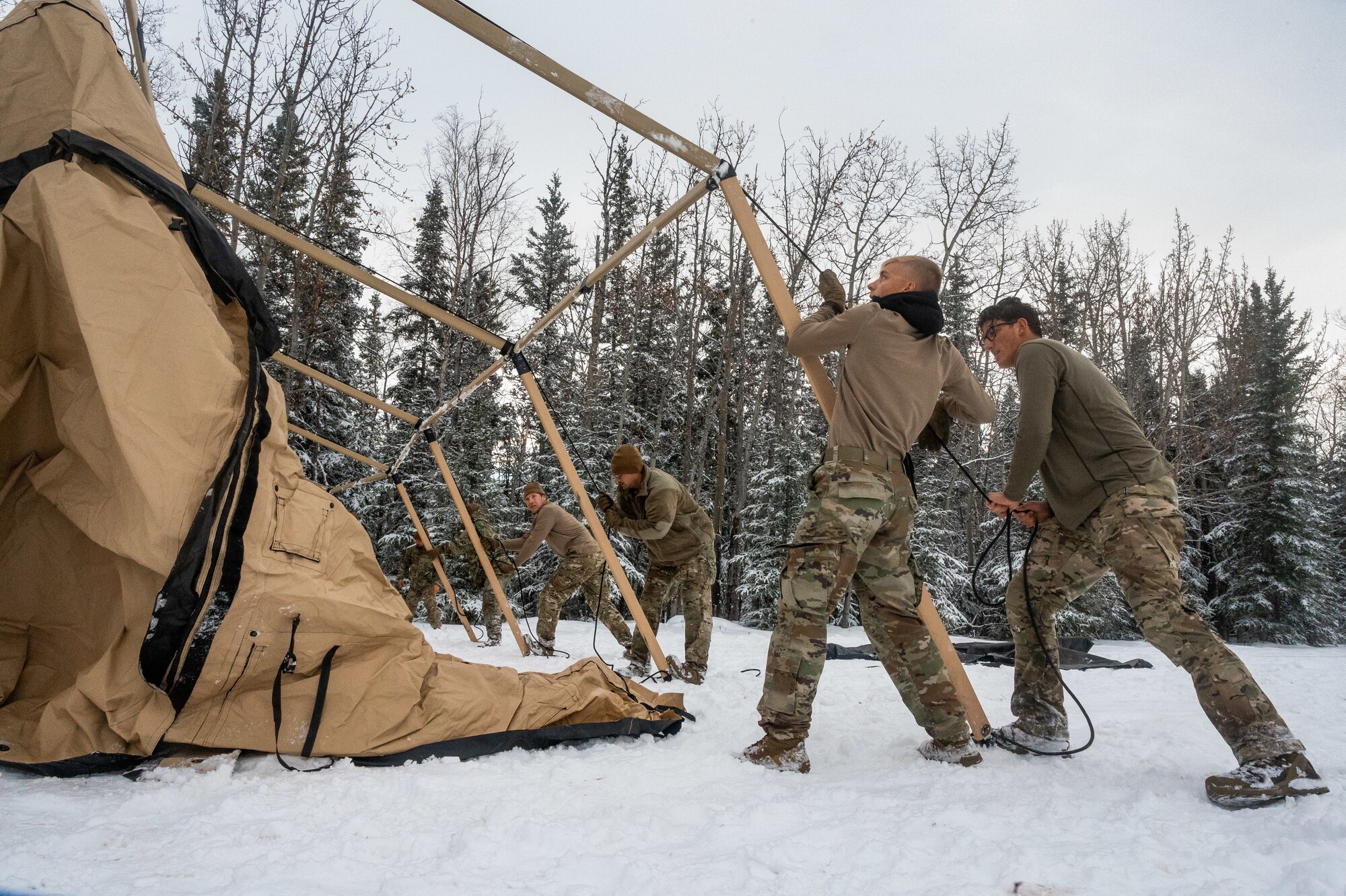 Six Airmen standing in snow pull a tent tarp up to the top of a tent frame with a rope in front a tree line,
