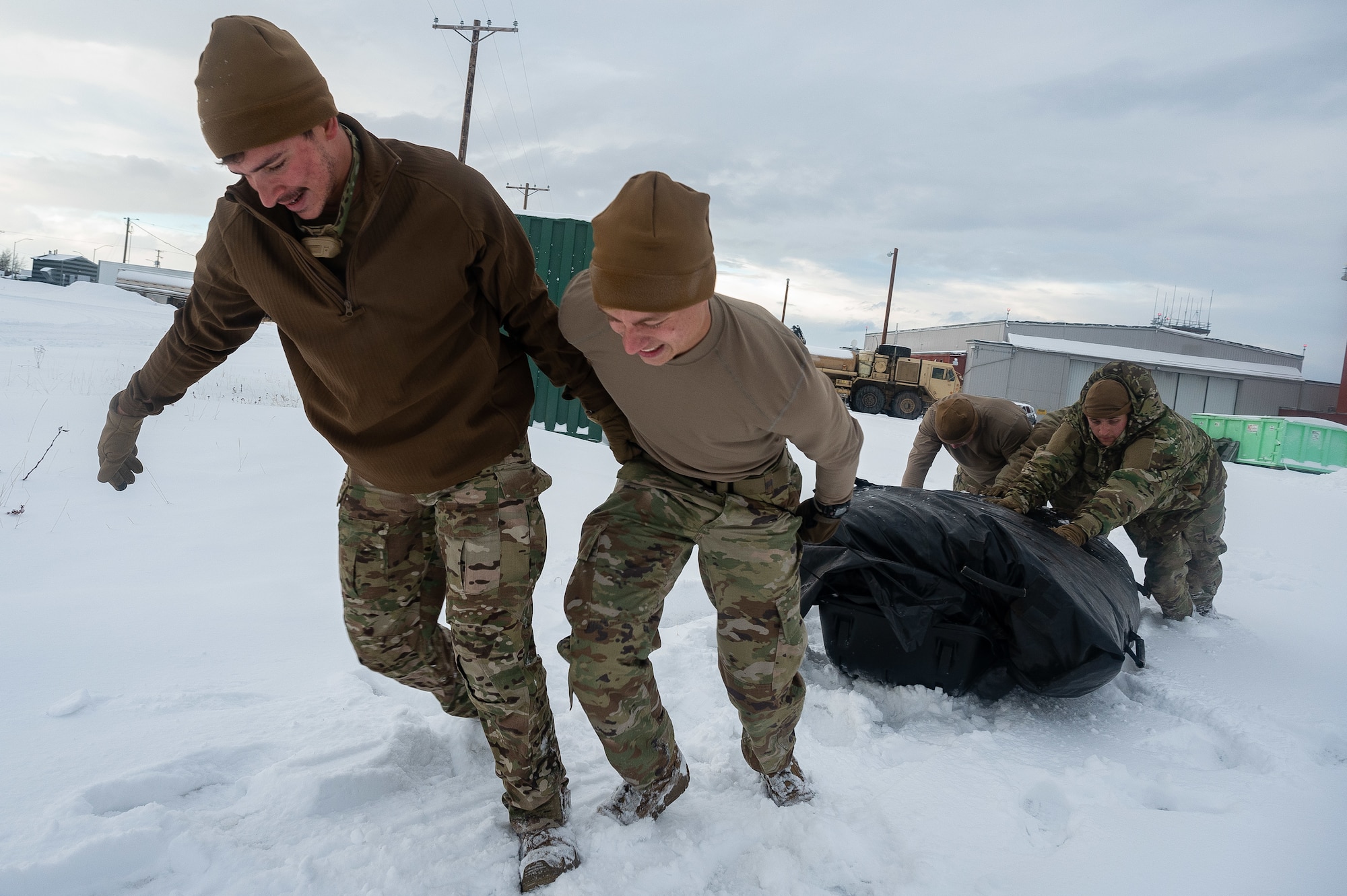 Two Airmen pull and two Airmen push a tarp through snow.