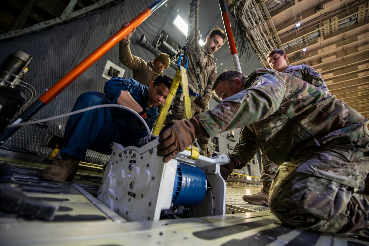 Men in military uniform replace a broken piece of machinery that has rope on it that is used to haul very large items on to very large military aircraft.