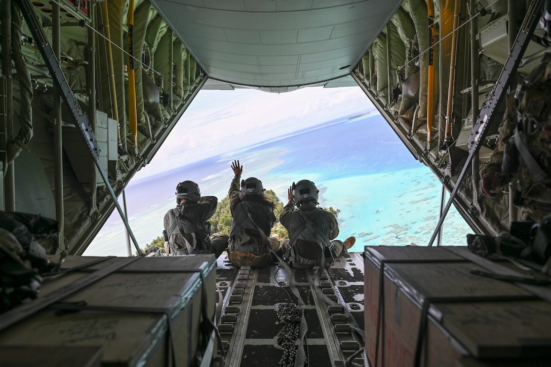 Three airmen sit at the back of an open aircraft flying over blue water; one waves.