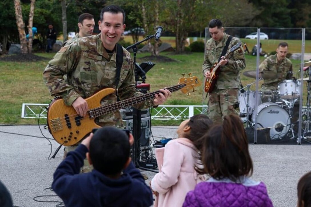 A bass guitarist in OCP uniform smiles as he performs for a crowd. Several children in colorful coats are dancing in the foreground. The rest of the band plays behind him on a stage set up in a grassy park with a few trees in the background.