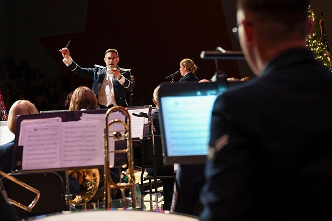 A photo taken from the backrow of the Heritage Band, featuring the commander conducting. The unit is wearing the AF mess dress, dominating colors are the blue of the uniform and the white of the music against a dark background.